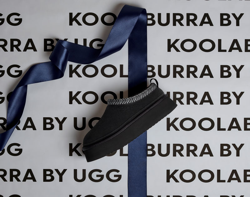 A Koolaburra slipper, laying on a blue ribbon and Koolaburra wrapping paper.